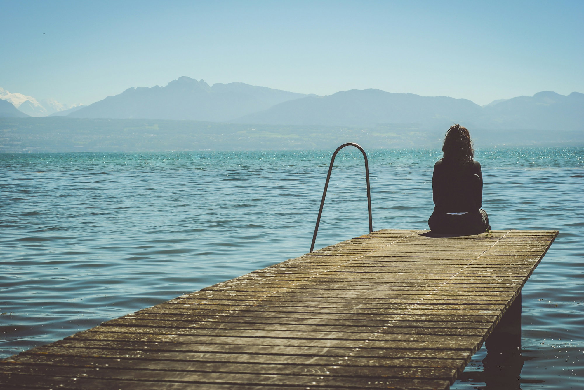 a-woman-sits-on-the-end-of-a-dock-during-daytime-staring-across-a-lake.jpg a woman sits on the end of a dock during daytime staring across a lake